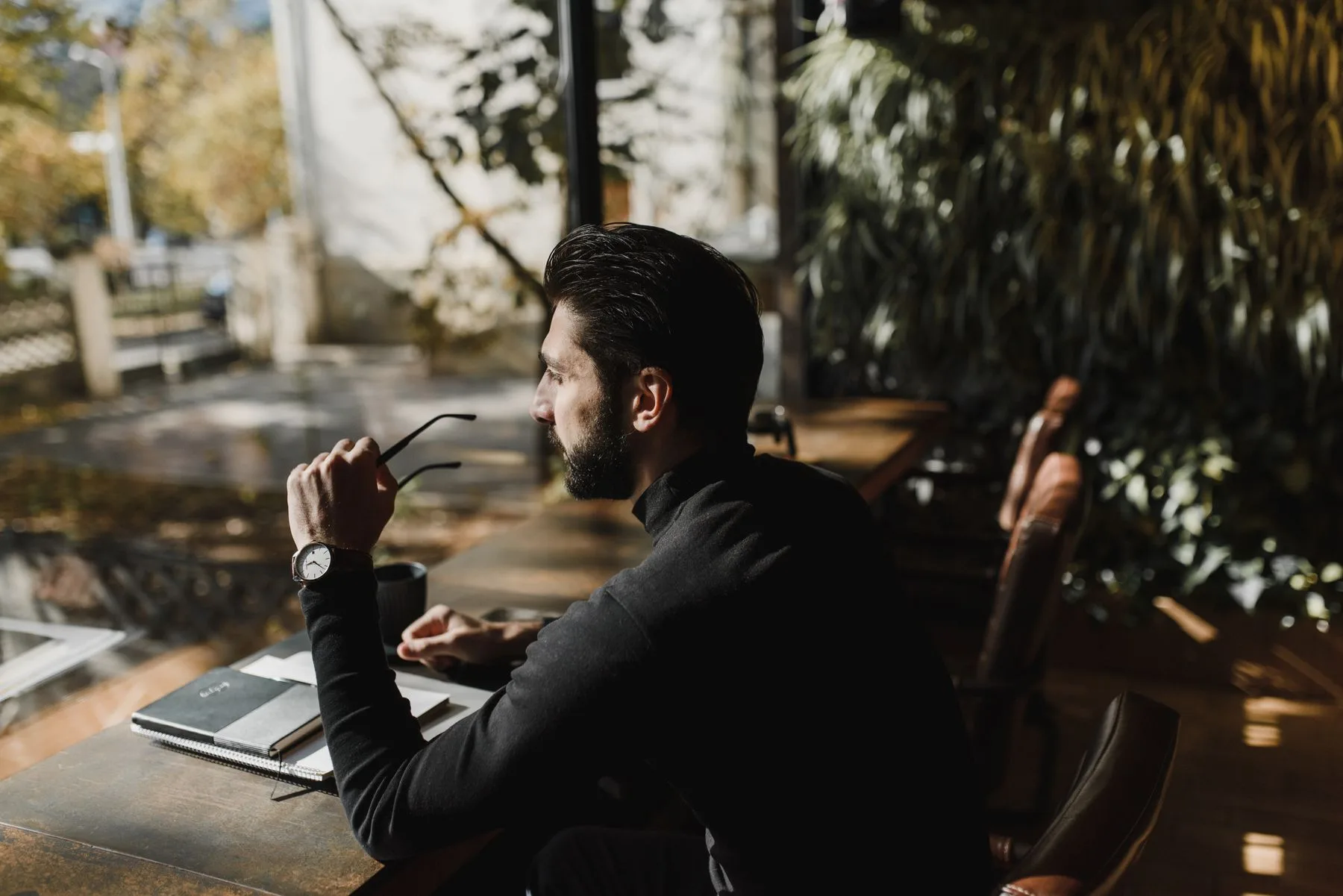 Man sitting by a sunlit window, lost in thought, holding glasses
